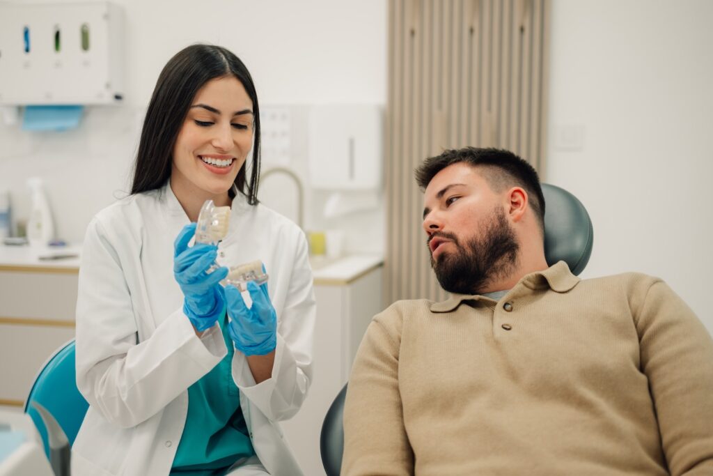 Female dentist wearing gloves and a lab coat smiling while showing a dental model to a male patient sitting in a dental chair, demonstrating a procedure or explaining oral health in a modern clinic.