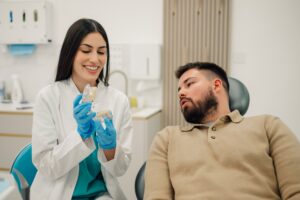 Female dentist wearing gloves and a lab coat smiling while showing a dental model to a male patient sitting in a dental chair, demonstrating a procedure or explaining oral health in a modern clinic.